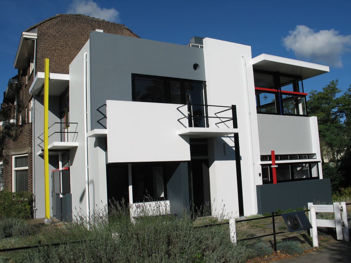 Das Rietveld Schröder Haus in Utrecht: weiße und graue Flächen, schwarze Fensterrahmen, ein gelber Pfosten sowie rote und blaue Stäbe unter blauem Himmel